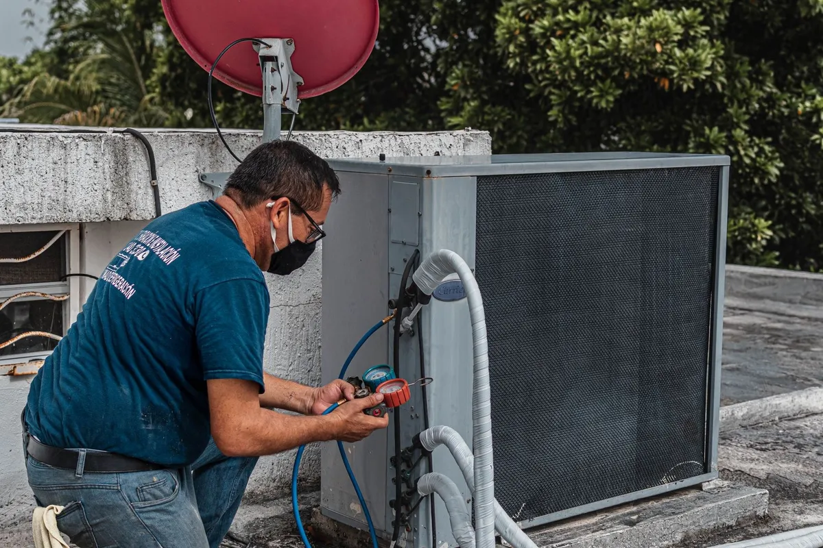A technician in a facemask repairs an outdoor air conditioner using a manifold gauge, illustrating HVAC maintenance.